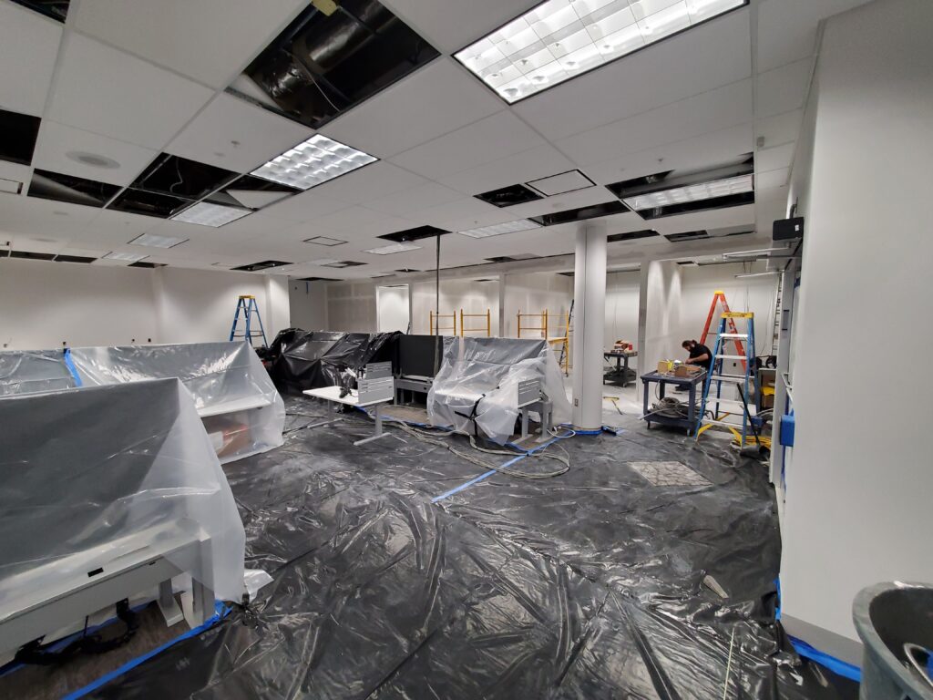 A room with desks and the floor covered in plastic. Wires hang from the drop ceiling. A person tends to wires in the corner.