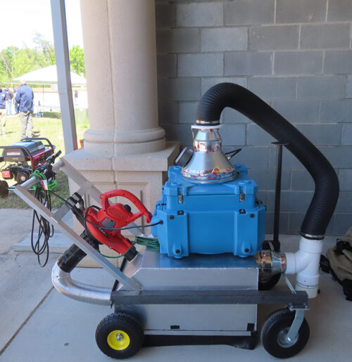 A machine with a push handle and wheels has a silver box at the bottom with a black tube connected to a smaller blue box at the top.