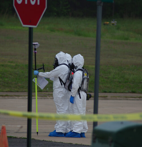 Three people in hazmat suits log samples with a device on a vertical pole.