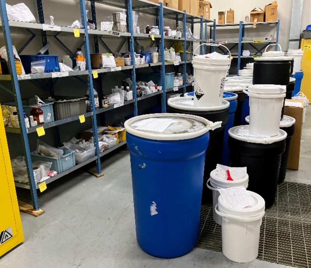Shelves organized with hazardous waste. Buckets and large drums line the center of the room.