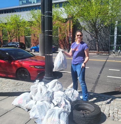 Woman stands on a sidewalk holding a bag of trash near a pile of trash bags.