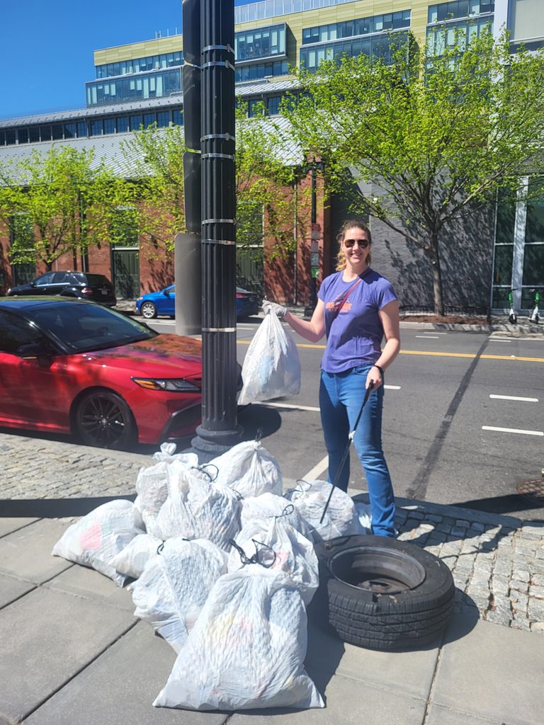 Woman stands on a sidewalk holding a bag of trash near a pile of trash bags.